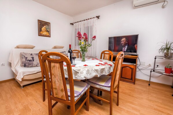 Un comedor con una mesa dispuesta para cuatro personas, sillas de madera, un jarrón con flores, un televisor en la pared y un sofá con cojines.