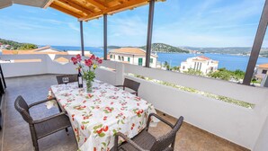 A covered outdoor patio with a table set for two, wicker chairs, and a view of the sea and buildings in the background. - Apartments Mirta (Razanj)