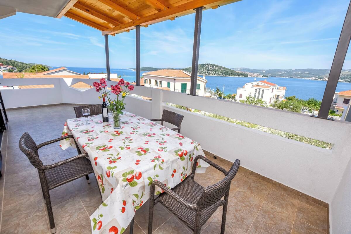 A covered outdoor patio with a table set for two, wicker chairs, and a view of the sea and buildings in the background.