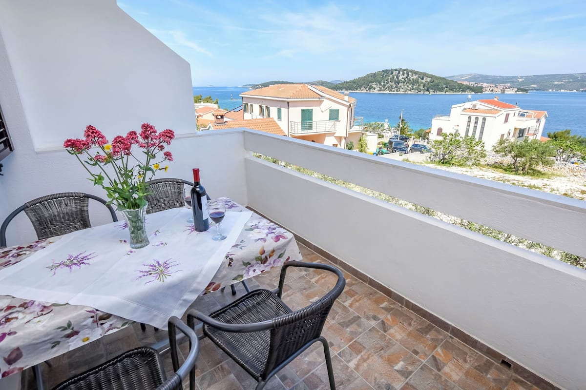 A balcony with a table set for two, a vase with flowers, and a view of the sea and buildings.