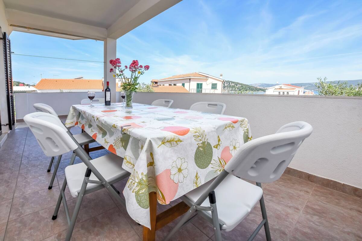 A covered outdoor dining area with a table set for two, white chairs, and a vase with flowers.