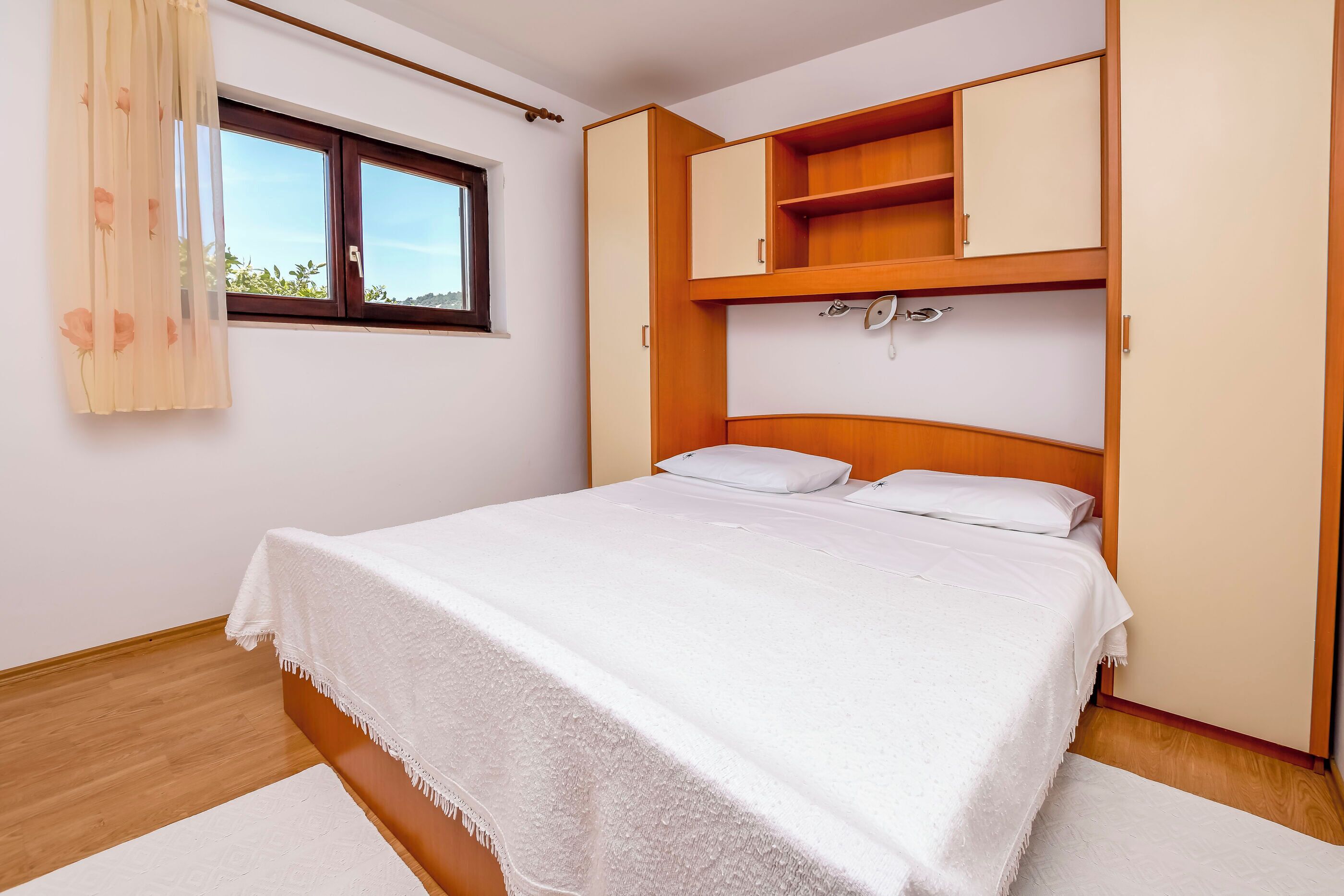 A bedroom with a bed, a window with curtains, and built-in wooden cabinets above the bed.