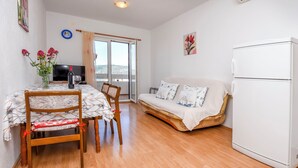 A modern living room with a wooden floor, a white sofa with patterned cushions, a dining table with chairs, a TV, and a refrigerator. - Apartments Mirta (Razanj)