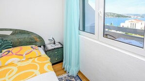 A bedroom with a bed, a nightstand, a lamp, a window with a view of a body of water and buildings, and a curtain. - Apartments Mirta (Razanj)