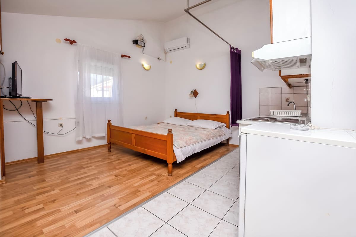 A bedroom with a wooden bed, a desk, a window with curtains, an air conditioner, and a kitchen area with a sink and a stove.