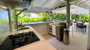 A kitchen with a gas stove, a sink, and a trash can. There is a dining area with chairs and a table. -  Viva Villa Village down the Fort  (Gosier)