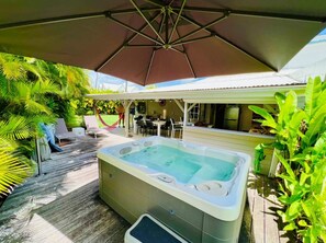 Un jacuzzi bajo un amplio paraguas, rodeado de mobiliario de exterior y abundante vegetación.