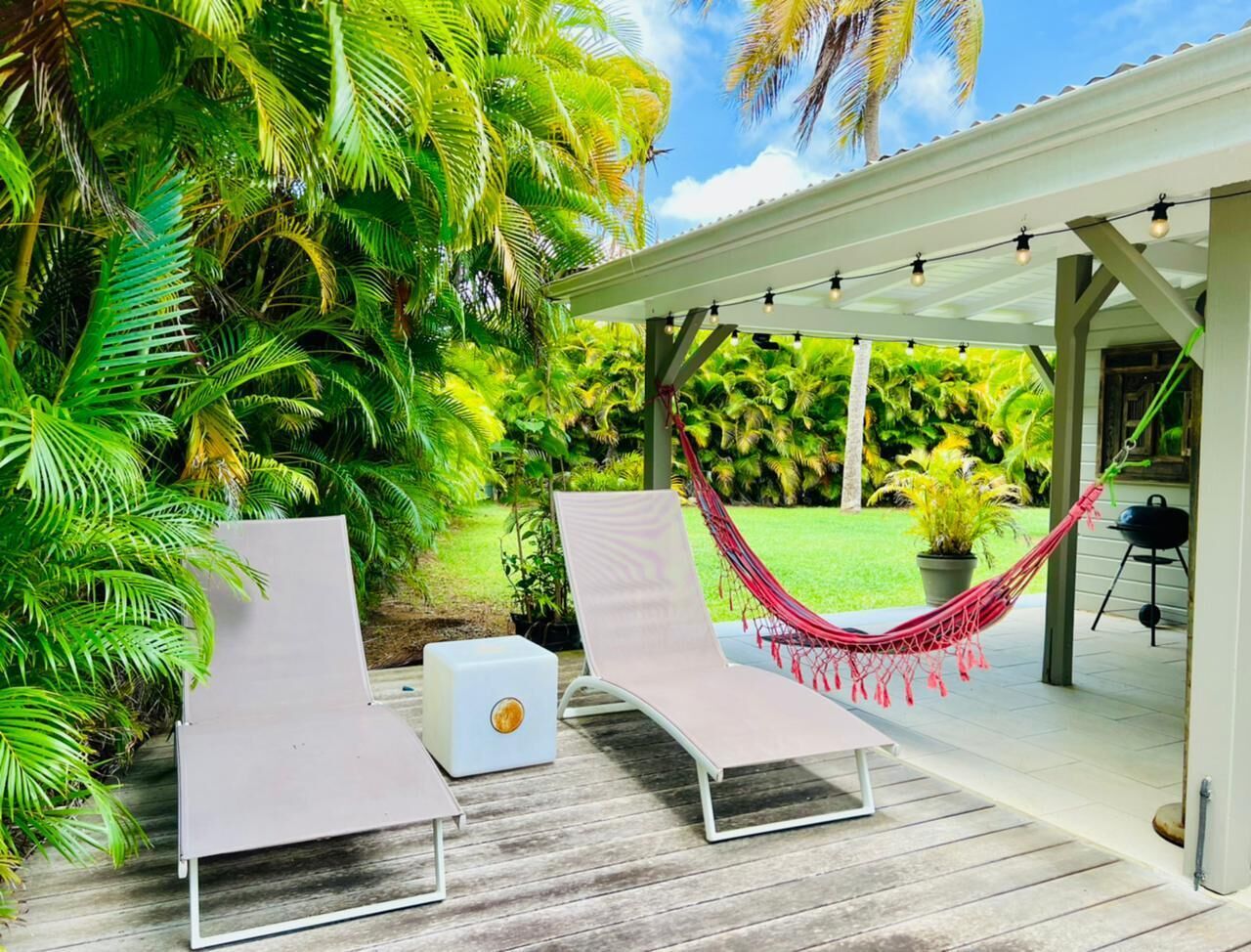 A covered outdoor area with two lounge chairs and a hammock, surrounded by lush tropical plants.