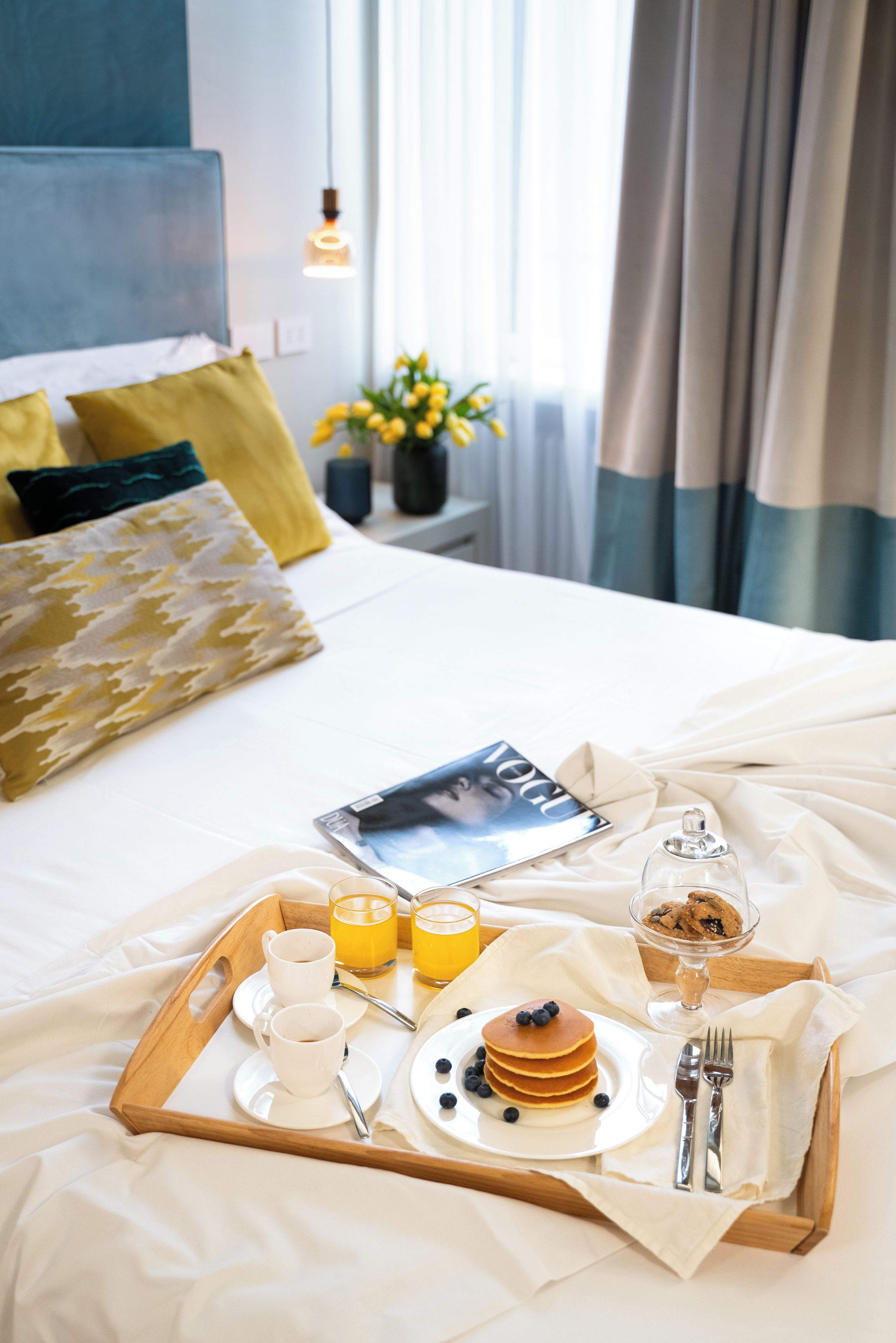 Una cama bien hecha con una bandeja de desayuno que incluye panqueques, café y jugo, en una habitación de hotel con vista a la ciudad.