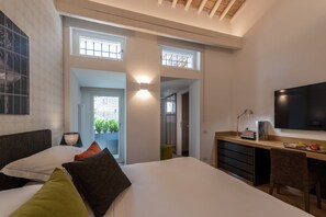 A modern bedroom with a bed, bedside table, and a desk area with a chair. There is a TV mounted on the wall and a window with a view of greenery outside. - Laurina 33 (Rome)