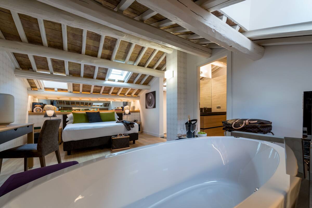 A spacious bedroom with a large white bathtub, a bed with a green and white headboard, and a wooden ceiling with skylights.