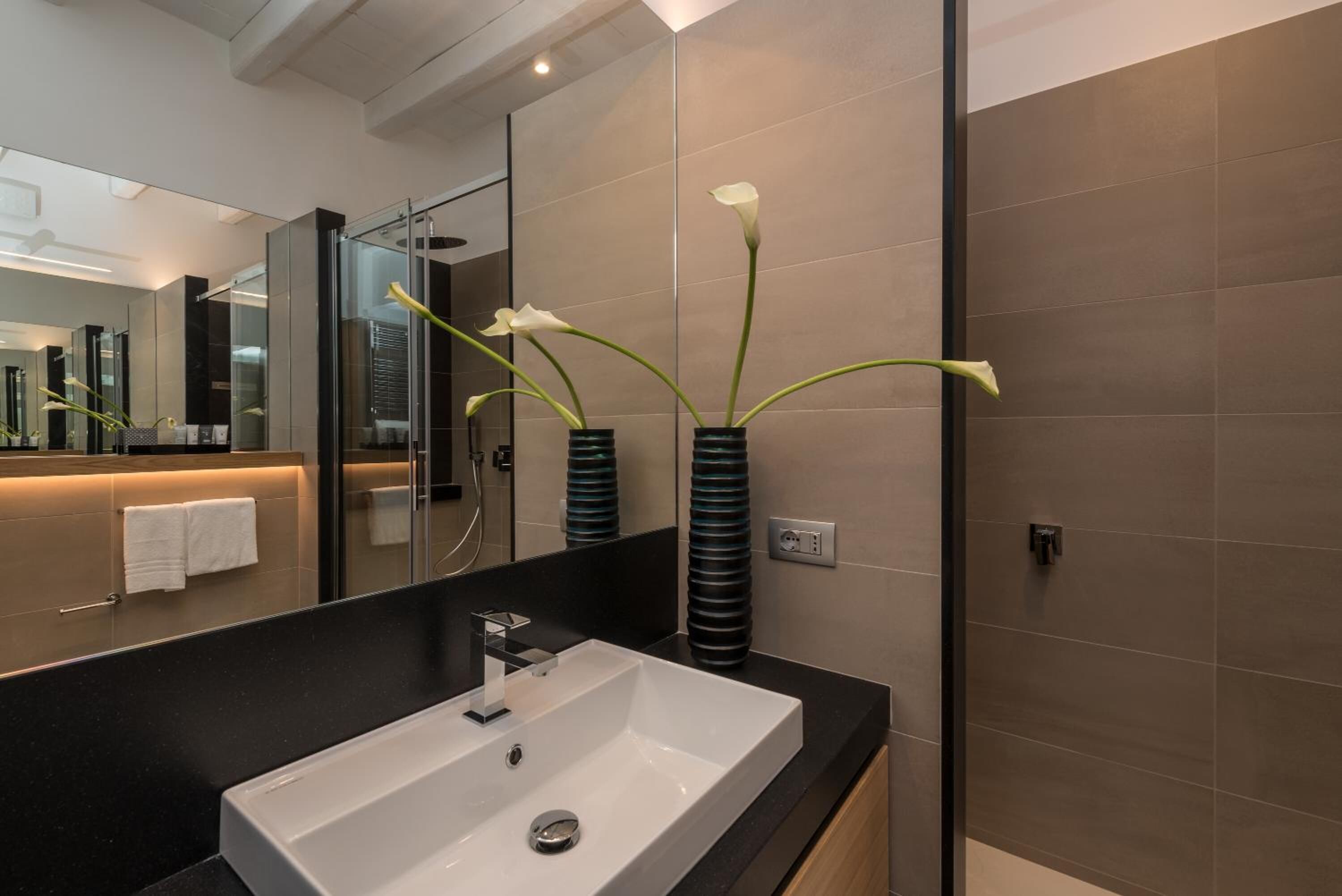 Un baño moderno con un espejo grande, un lavamanos blanco y altas jarrones negros con flores blancas.