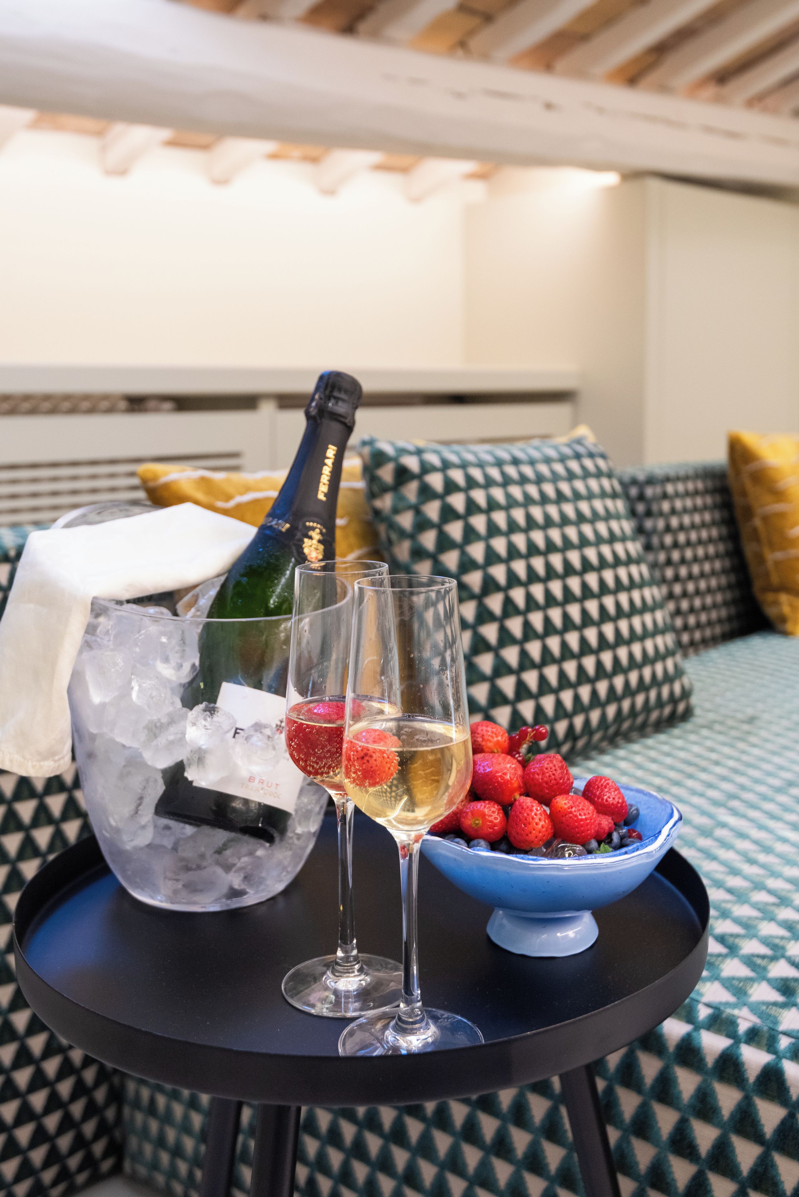 A bed with a geometric patterned bedspread, a bedside table with a bottle of champagne, glasses, and a bowl of berries.
