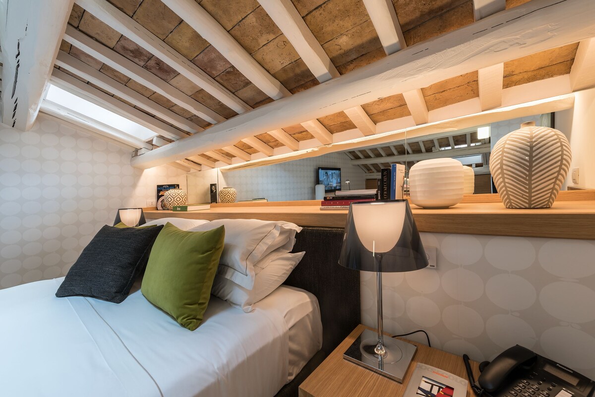 A modern hotel room with a wooden ceiling, a bed with white and green pillows, a bedside lamp, a nightstand with a phone, and a mirror above the bed.