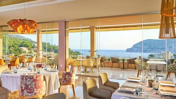 Ein Restaurant mit gedeckten Tischen, großen Fenstern mit Blick auf Meer und Hügel sowie moderner Beleuchtung.