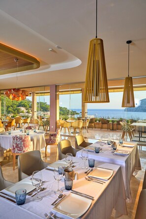 A restaurant with tables set for dining, large windows offering a view of the sea, and hanging wooden pendant lights. - Zafiro Palace Andratx (Andraitx)