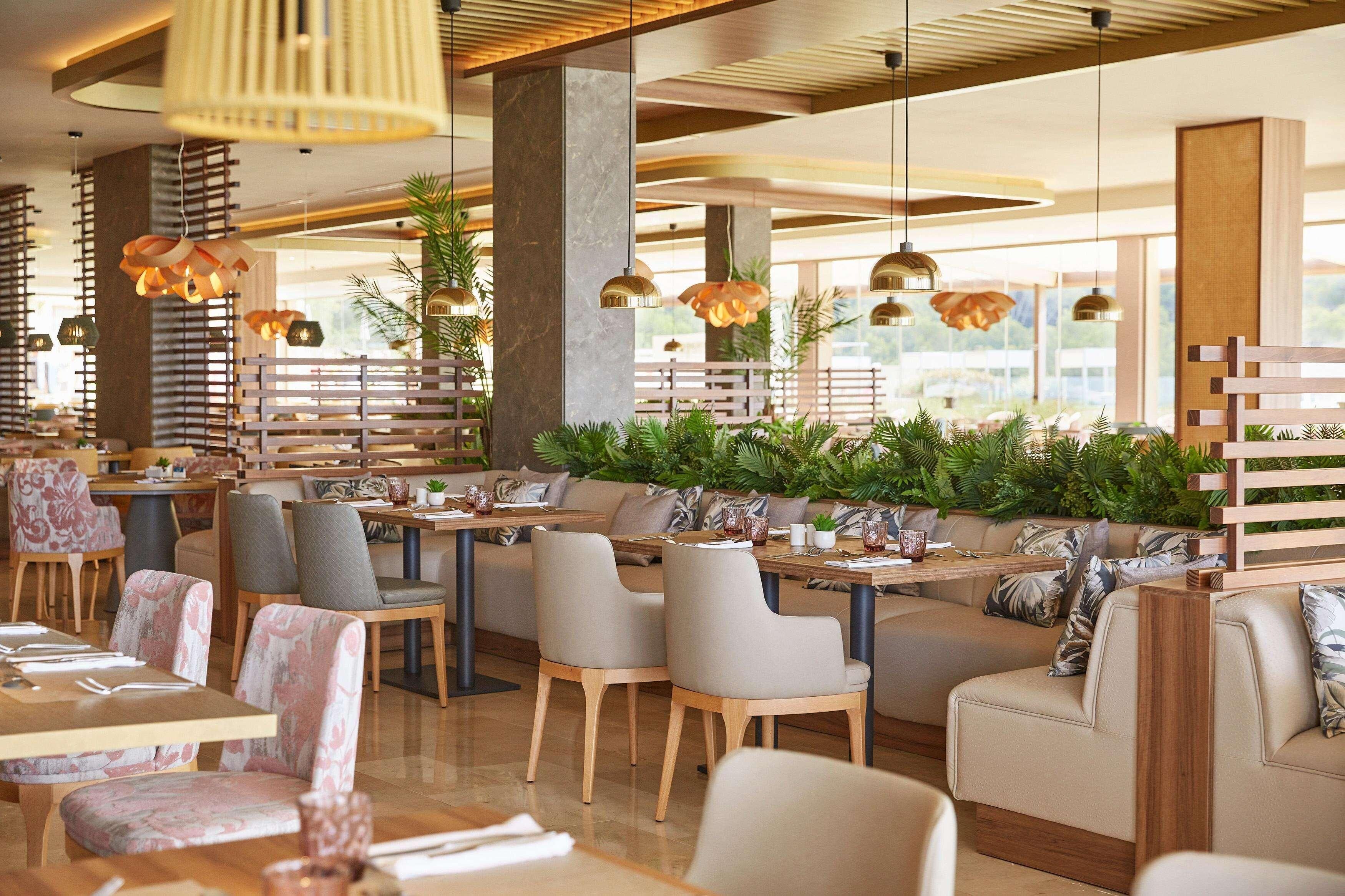 Un restaurante moderno con mesas y sillas de madera, grandes lámparas colgantes y una zona de estar con un banco de madera y cojines.