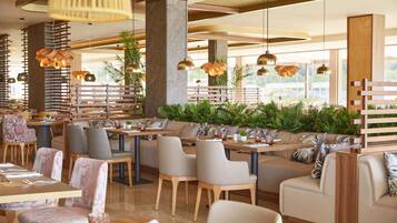 Un restaurante moderno con mesas y sillas de madera, grandes lámparas colgantes y una zona de estar con un banco de madera y cojines.
