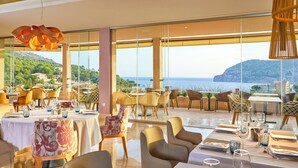 Un restaurante con mesas dispuestas para comer, amplios ventanales que ofrecen vistas al mar y a las colinas, y lámparas de diseño moderno.