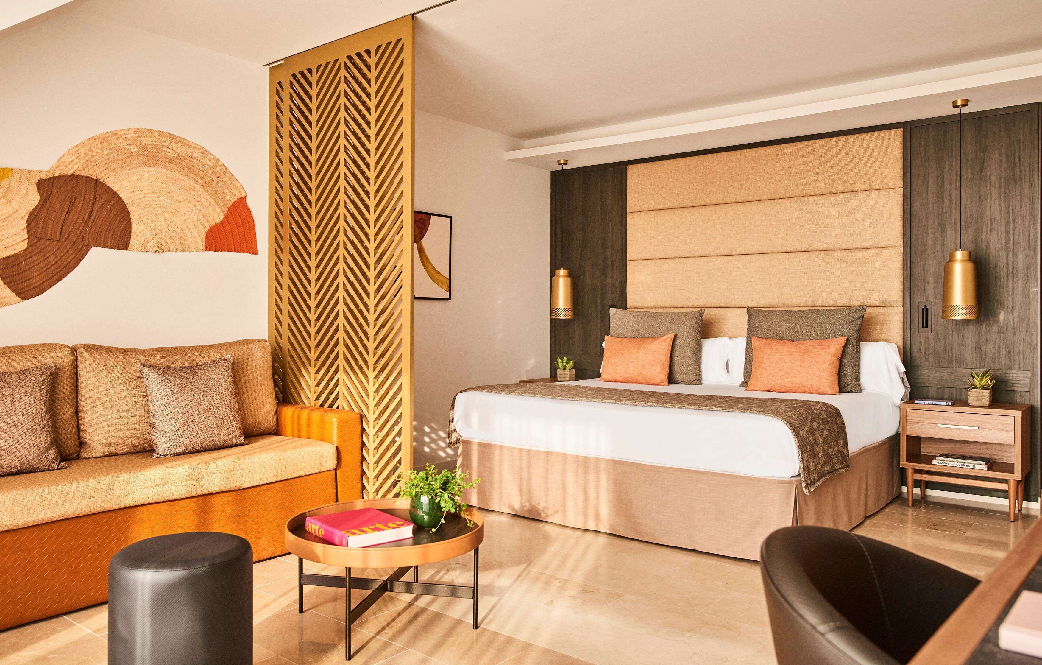 Een moderne hotelkamer met een bank, een bed met veel kussens, een nachtkastje met een lamp en een decoratief wandpaneel.