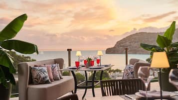 A dining area with a table set for two, overlooking a scenic view of the sea and hills.