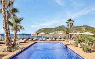 A pool area with lounge chairs and umbrellas, surrounded by palm trees and a view of the sea and hills.