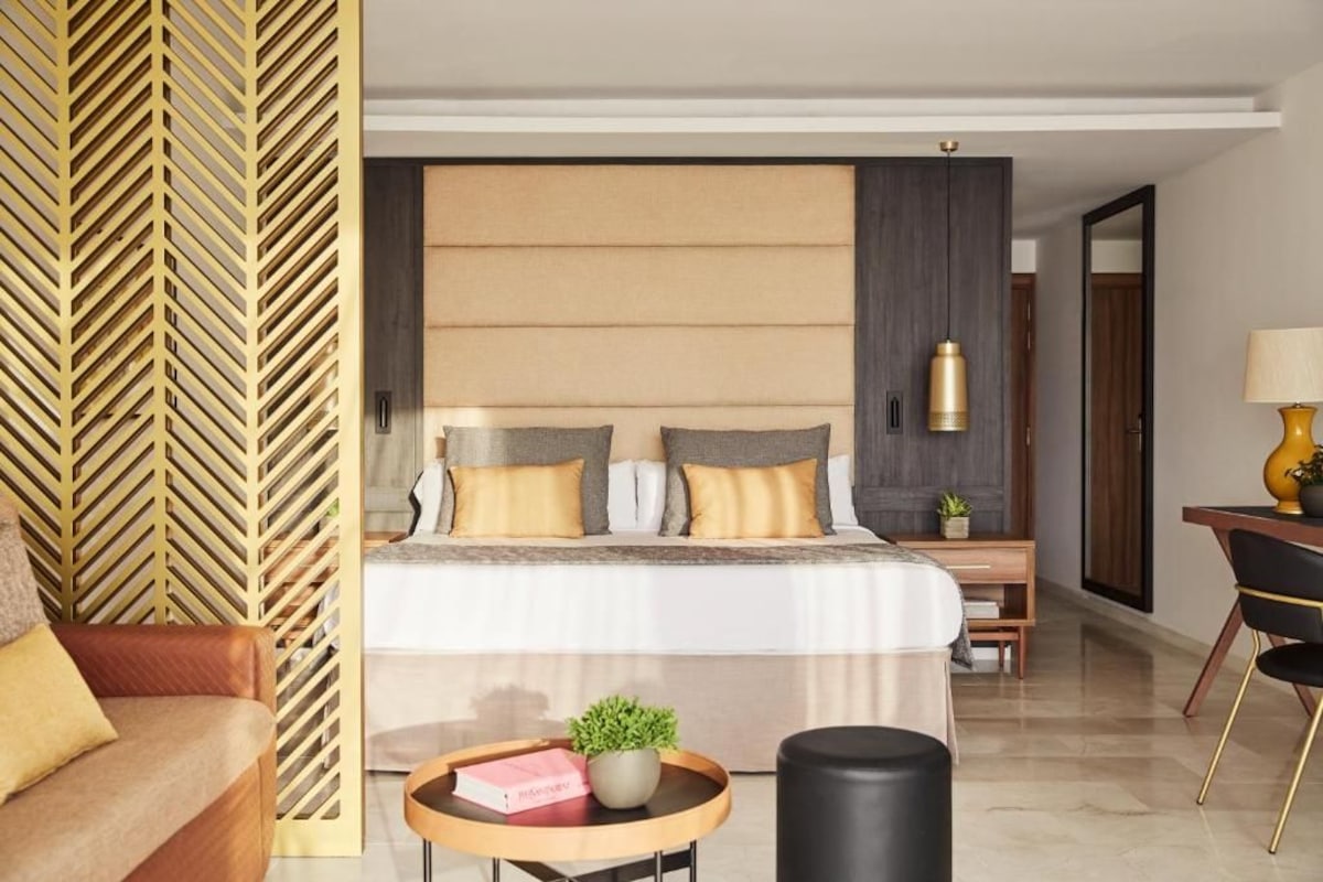 Une chambre d’hôtel moderne avec un grand lit, une tête de lit en bois, une petite table avec une plante et un bureau avec une lampe.