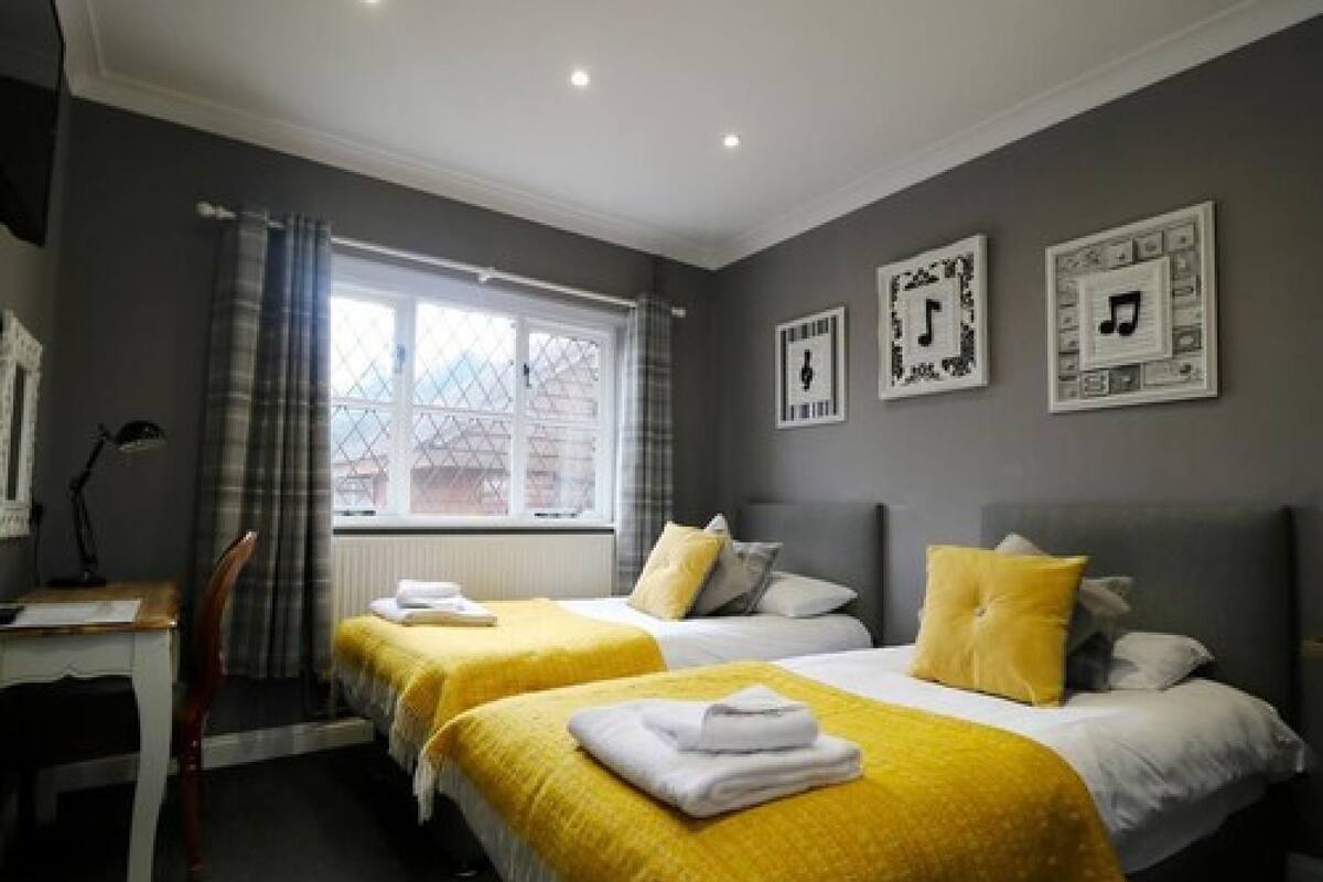 Quarto de hotel com duas camas de solteiro, uma escrivaninha, uma cadeira e uma janela com cortinas.