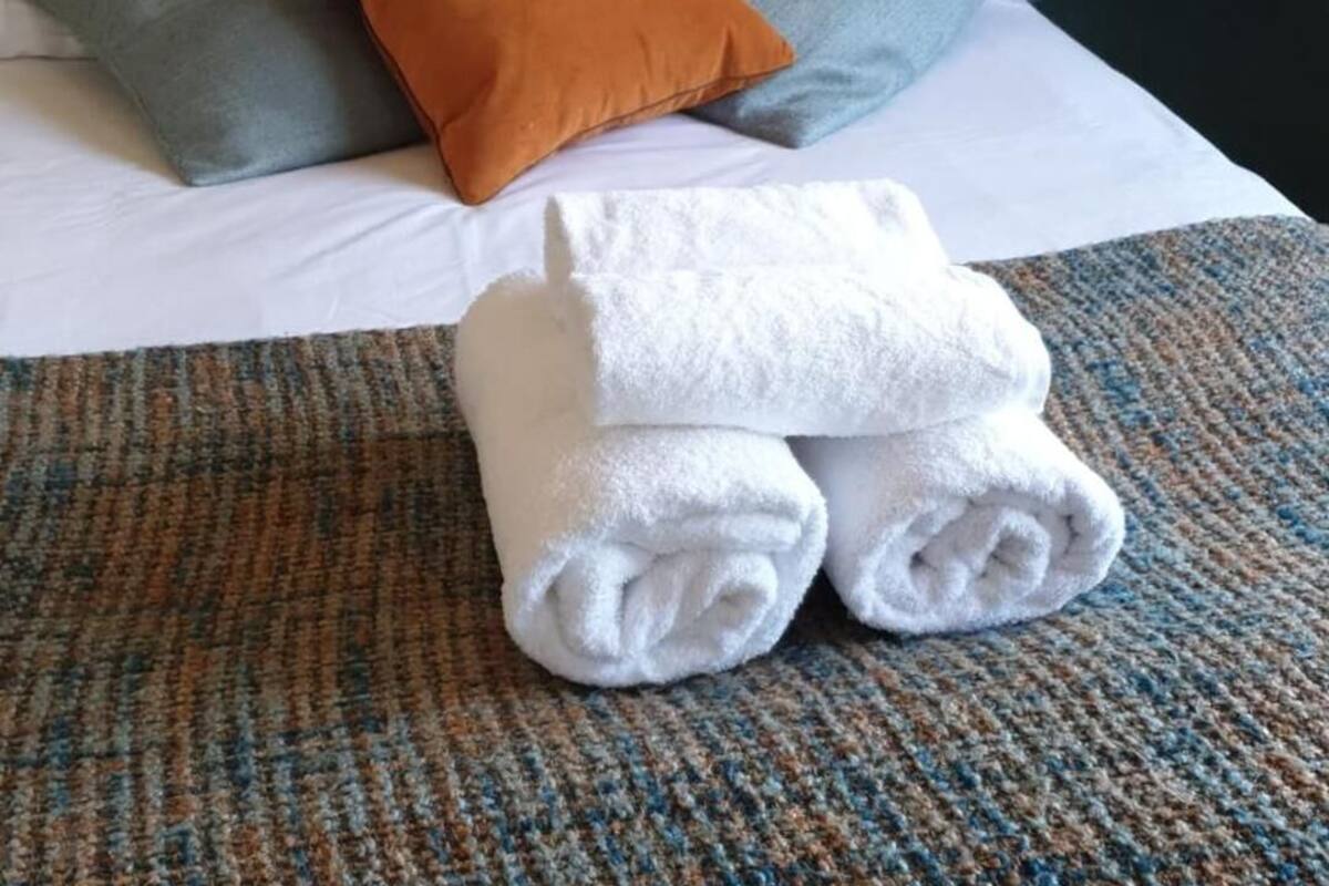 Cama bem arrumada com almofadas e toalhas dobradas sobre um banquinho com textura.