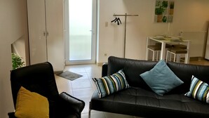 Moderni olohuone, jossa on musta nahkasohva, raidallisia koristetyynyjä ja keltainen heittotyyny. Olohuoneessa on myös ruokailutila, jossa on valkoinen pöytä ja tuolit, sekä naulakko, jossa on vaatteita. Liukuovet johtavat parvekkeelle.