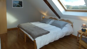 Et soveværelse med skrå vægge, en seng med gråt og hvidt dynebetræk, et lille natbord med en lampe og et billede på væggen.
