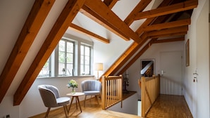 Ein Schlafzimmer im Loft-Stil mit freiliegenden Holzbalken, einer Holztreppe, einer kleinen Sitzecke mit Sessel und Tisch und einem Fenster, das für natürliches Licht sorgt.