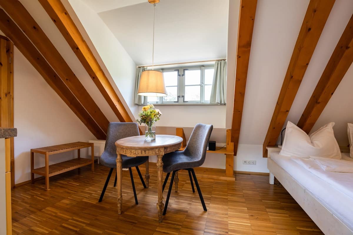 Ein gemütliches Schlafzimmer im Dachgeschoss mit Holzboden, einem runden Esstisch mit Stühlen, einem kleinen Fenster mit Vorhängen und einem Bett mit weißer Bettwäsche.