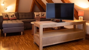 Ein gemütliches Dachzimmer mit Holzbalkendecke, einem Flachbildfernseher auf einem Holzgestell, einer Couch und einem kleinen Tisch mit einer Lampe.