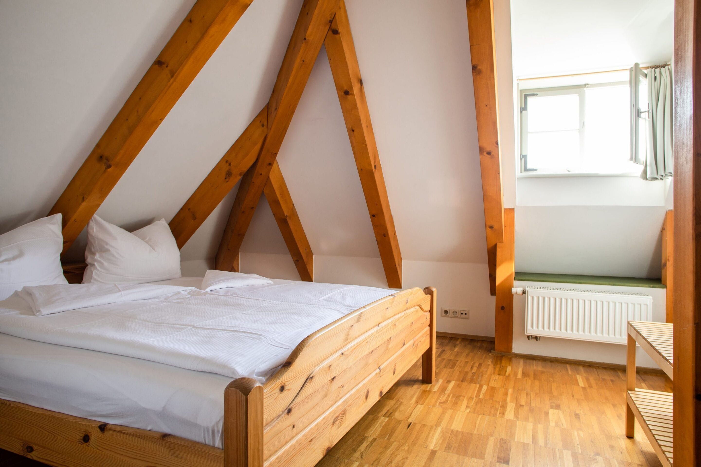 Quarto com cama de madeira, roupa de cama branca e teto inclinado com vigas de madeira.