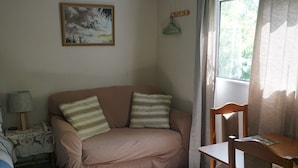A room with a sofa, a wooden chair, a small table, a lamp, and a painting on the wall. - Stand alone cottage in tranquil garden, with balcony, pergola, SkyTV, free Wifi (Greytown)