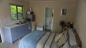 A bedroom with a bed, a nightstand, a desk with a sink, and a window with blinds. - Stand alone cottage in tranquil garden, with balcony, pergola, SkyTV, free Wifi (Greytown)