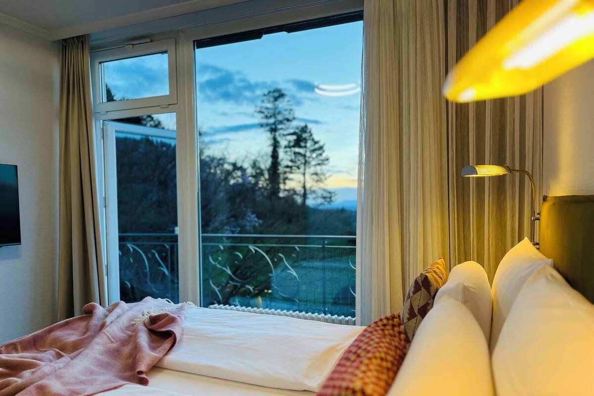A hotel room with a large window, a bed with white linens, a pink blanket, and a patterned throw.