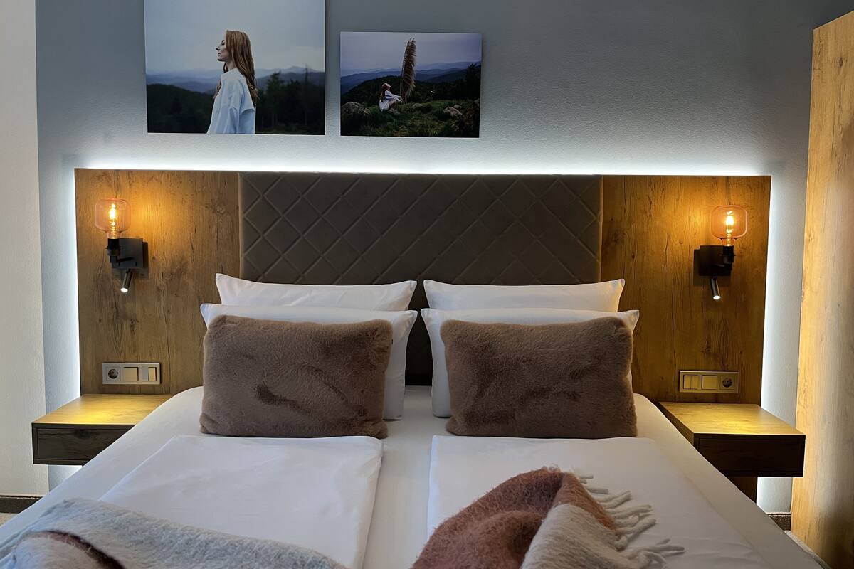 A double bed with white and brown bedding, two wall-mounted light fixtures, and two framed pictures above the bed.