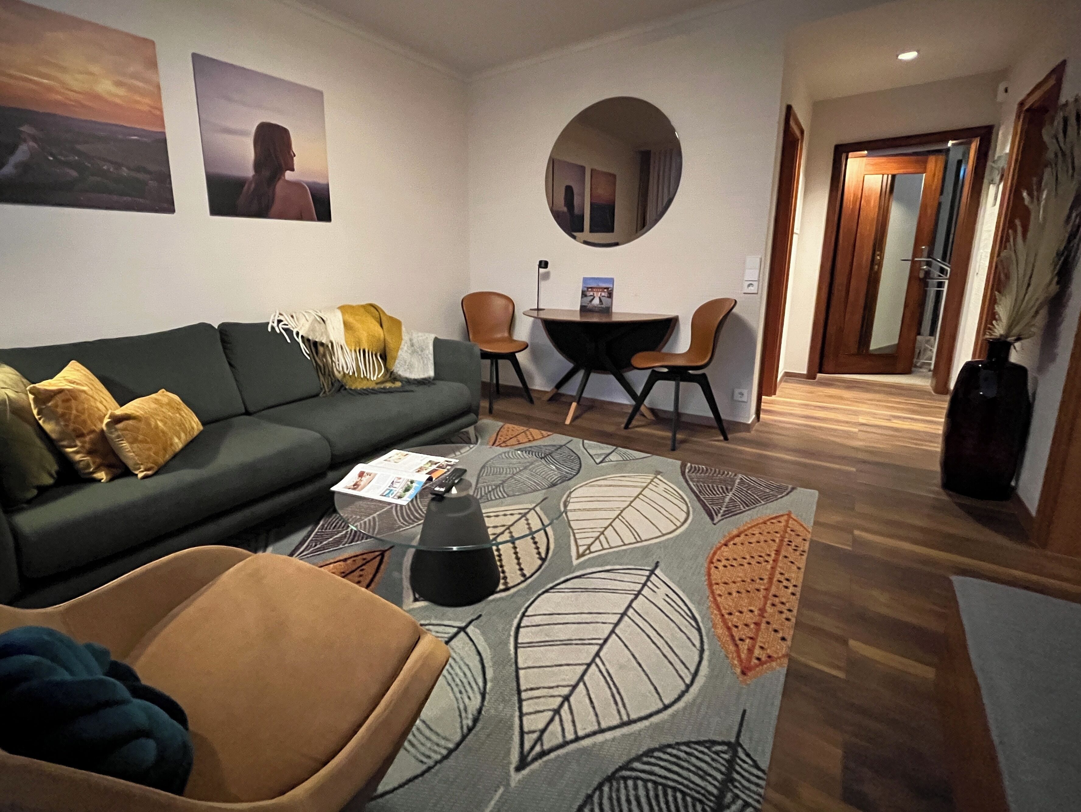A living room with a sofa, a coffee table, a round mirror, and a dining area with chairs.