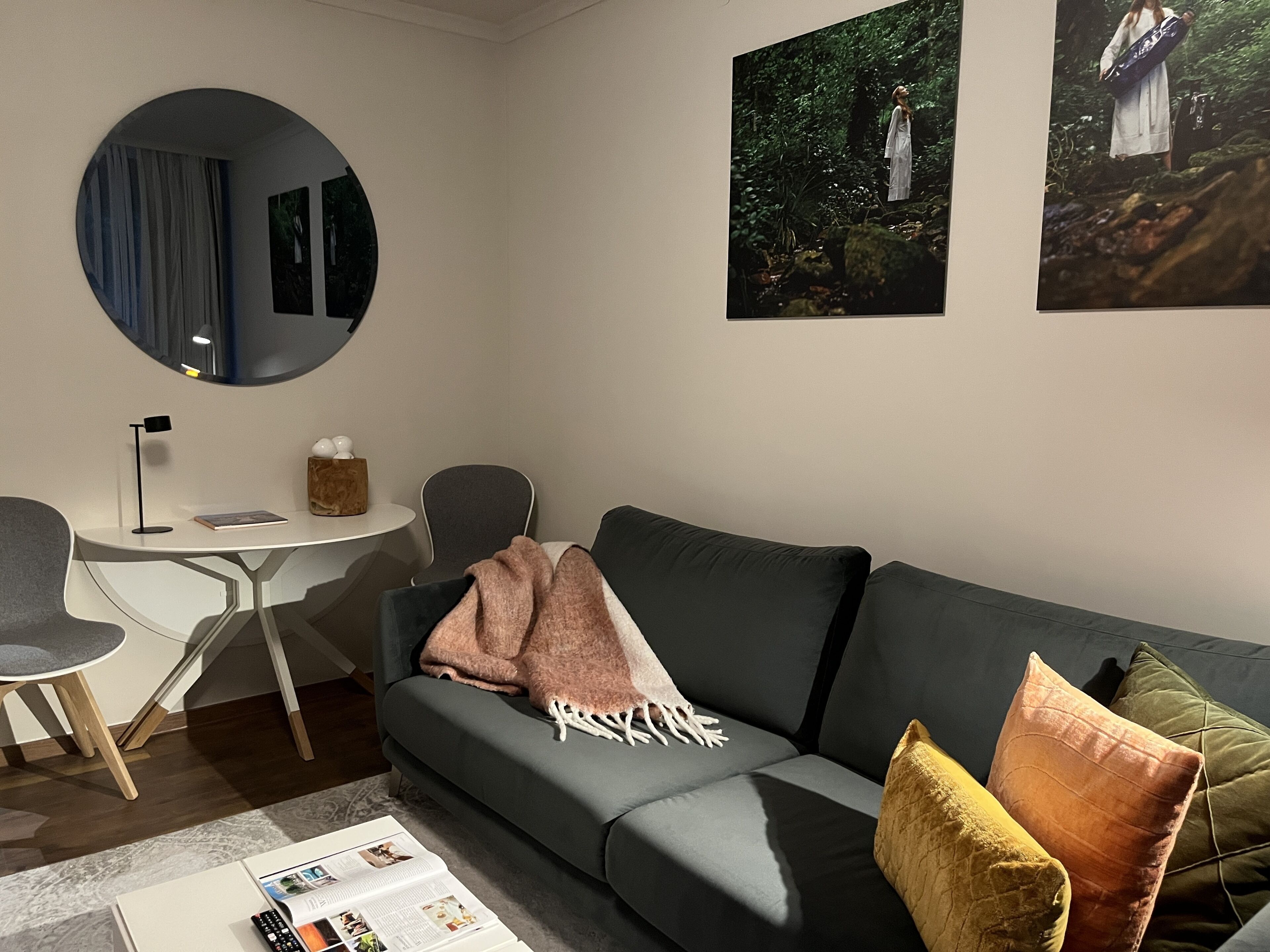 Ein modernes Wohnzimmer mit einem grauen Sofa, einem runden Spiegel, einem kleinen runden Tisch und einem Sessel. An der Wand befinden sich zwei gerahmte Bilder.