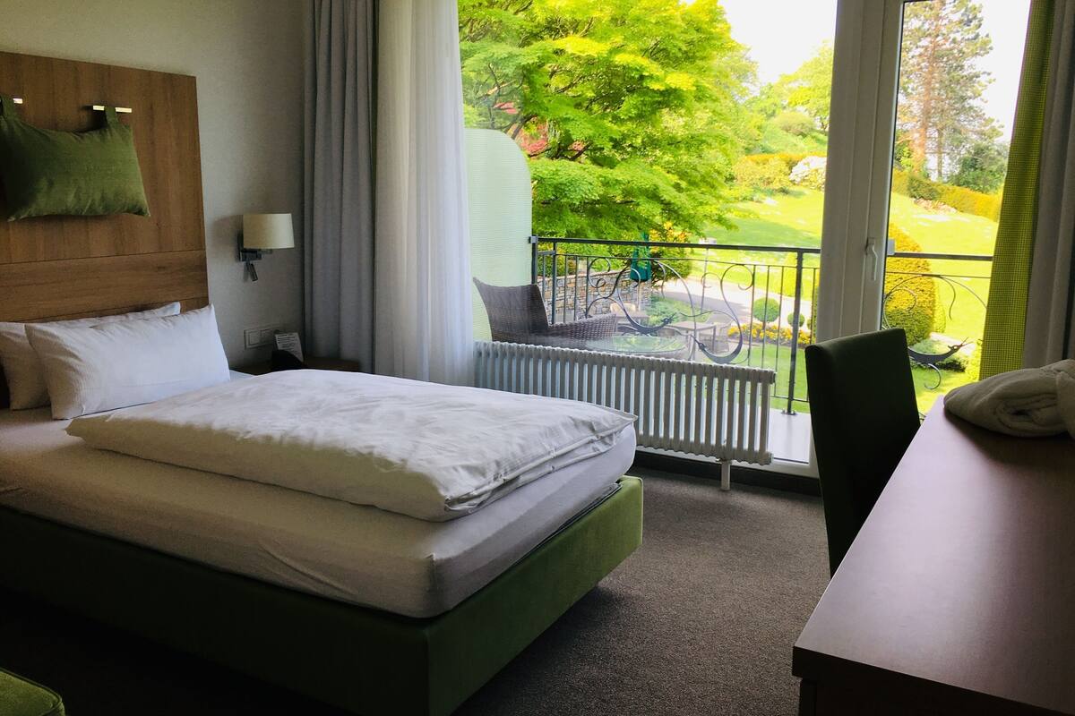 A hotel room with a bed, a table, a chair, a view of a garden through a window, and a balcony with a chair and a table.