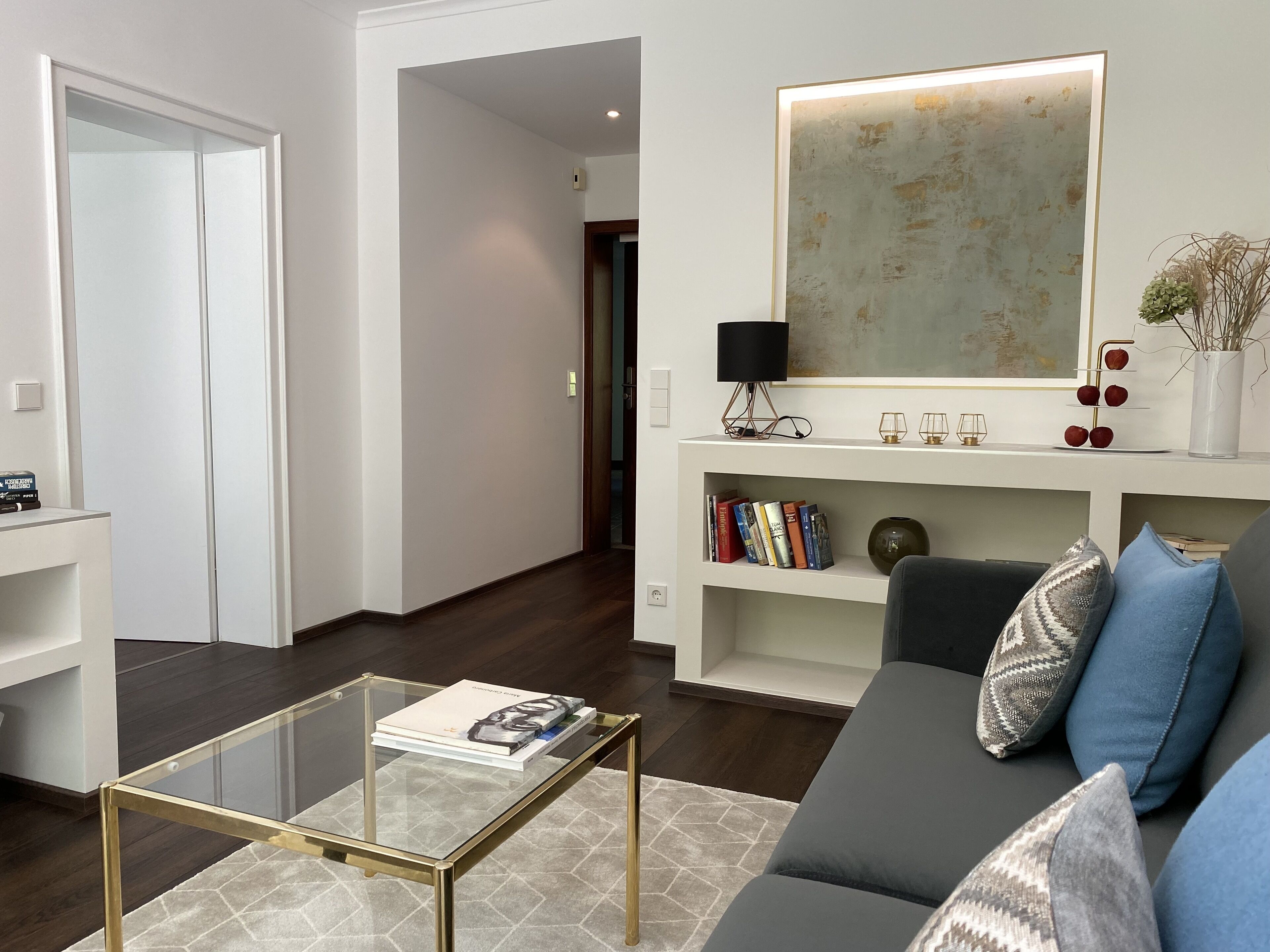 Ein modernes Wohnzimmer mit einem Glastisch, einem grauen Sofa mit gemusterten Kissen, einem Bücherregal mit Büchern und Dekoration sowie einem großen abstrakten Gemälde an der Wand.