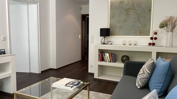 Ein modernes Wohnzimmer mit einem Glastisch, einem grauen Sofa mit gemusterten Kissen, einem Bücherregal mit Büchern und Dekoration sowie einem großen abstrakten Gemälde an der Wand.