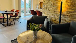 A modern living room with a stone accent wall, a dining area with red chairs, and a view through large glass doors. - Hotel Schlossberg (Badenweiler)