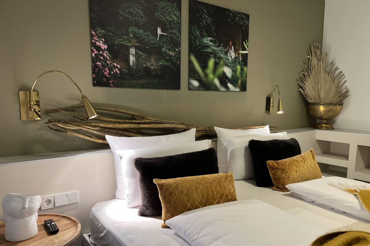 A hotel room with two beds, white bedding, a wooden side table with a remote control, a wall-mounted lamp, and two framed nature photographs.