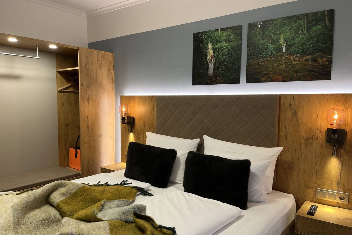 A modern hotel room with a bed, two wall-mounted lights, a nightstand, a wooden wardrobe, and two framed pictures on the wall.
