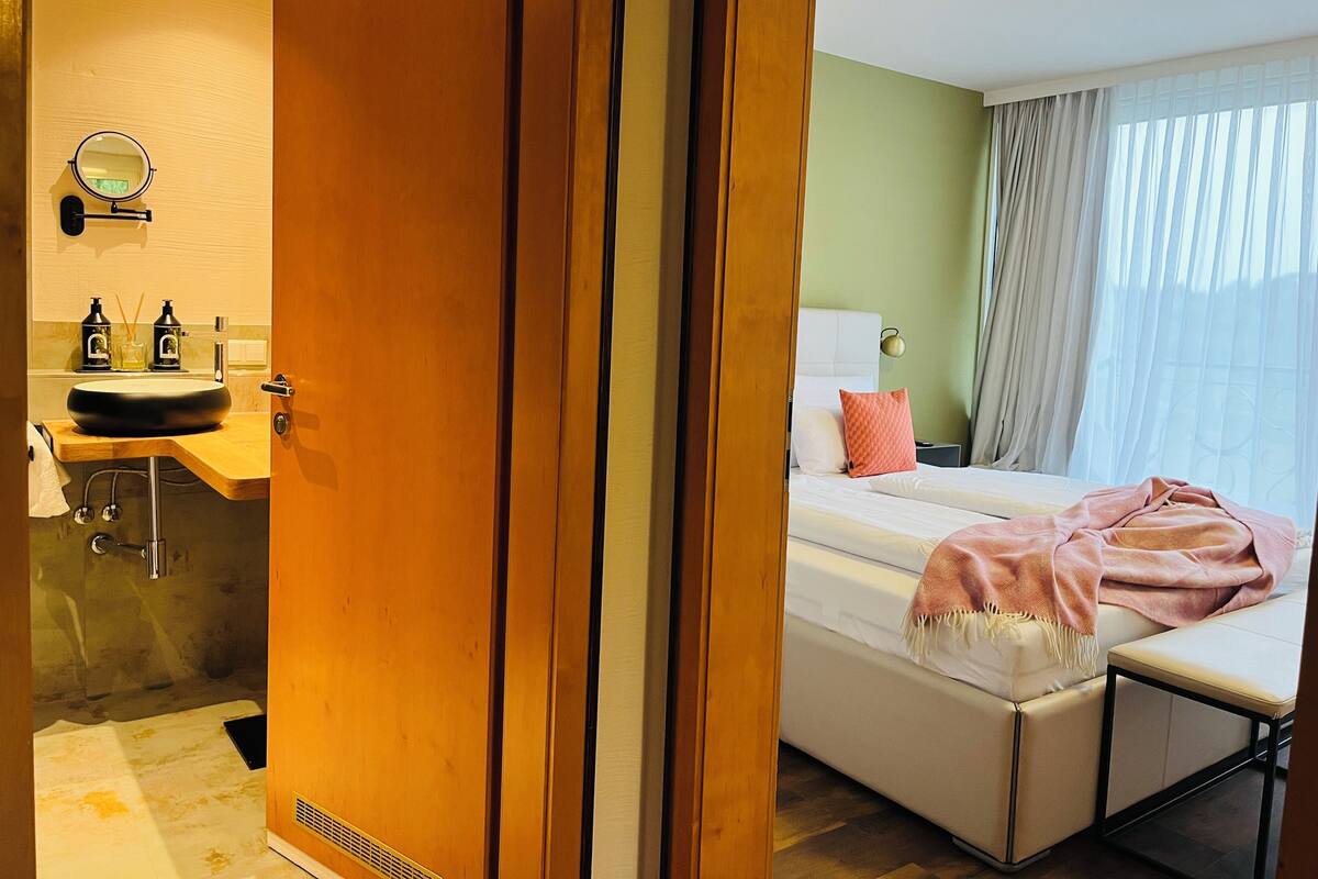 A hotel room with a wooden door, a sink with soap dispensers, a bed with a pink blanket, and a bedside lamp.