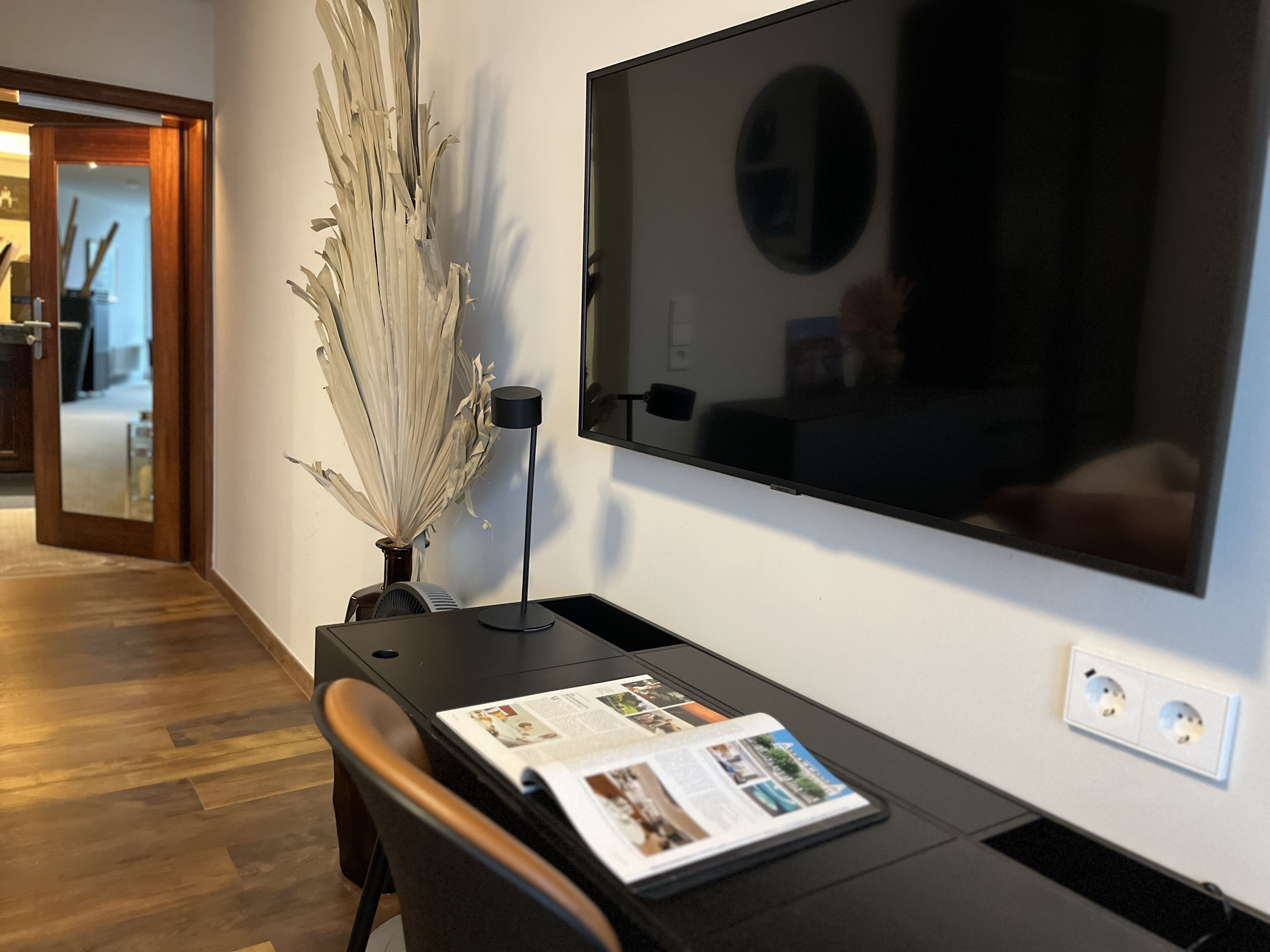 Una habitación moderna con un televisor de pantalla plana grande montado en la pared, suelo de madera, un escritorio con una revista y un jarrón decorativo con plantas secas.