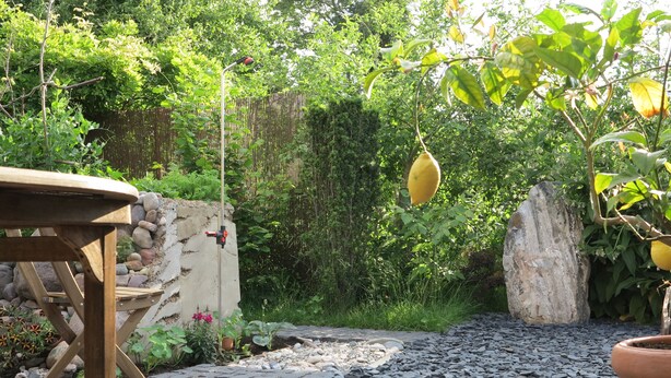 Um jardim com mesa e cadeiras de madeira, um muro de pedra e diversas plantas e árvores.